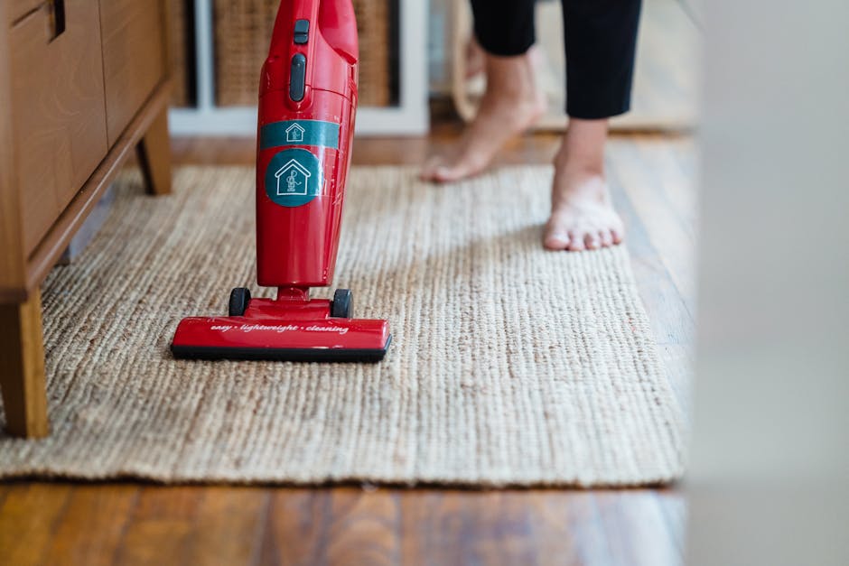A close-up of a vacuum cleaner with a clear dust container filled with debris, connected to a flexible, black and white patterned hose. The vacuum is positioned on a light pink carpet in a room with a