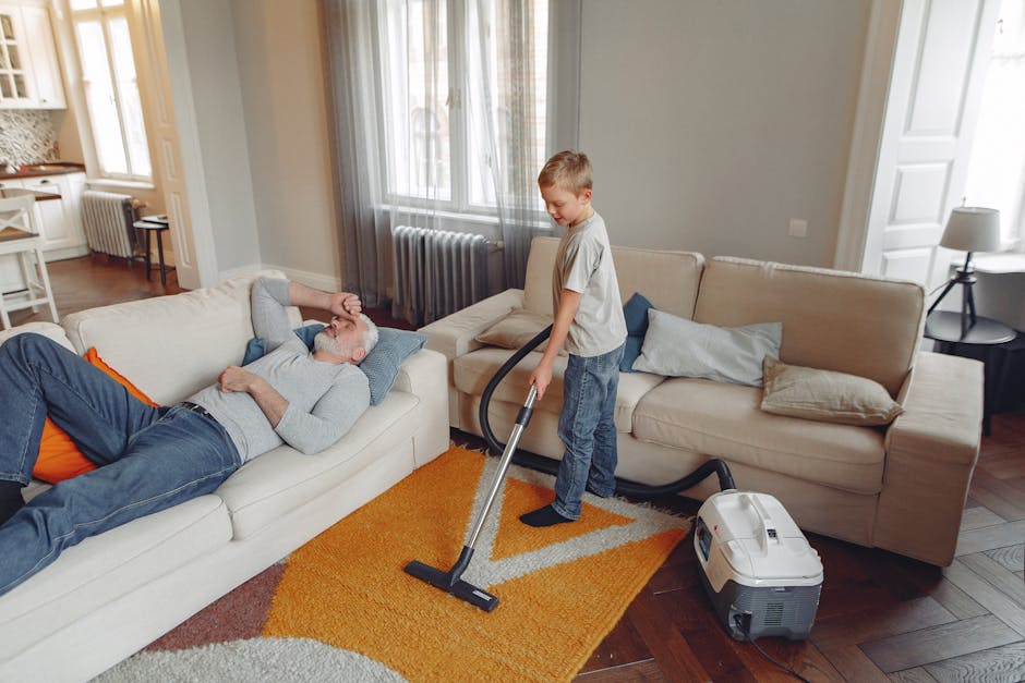 A person dressed in full white protective PPE, including a suit, gloves, face mask, and shoe covers, is performing deep cleaning on a beige carpet with a large industrial vacuum cleaner in a bright, m
